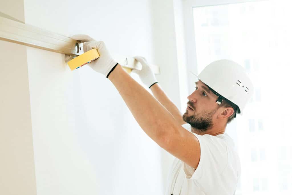 Contractor wearing a hard hat and gloves, measuring a wall for home improvement work.